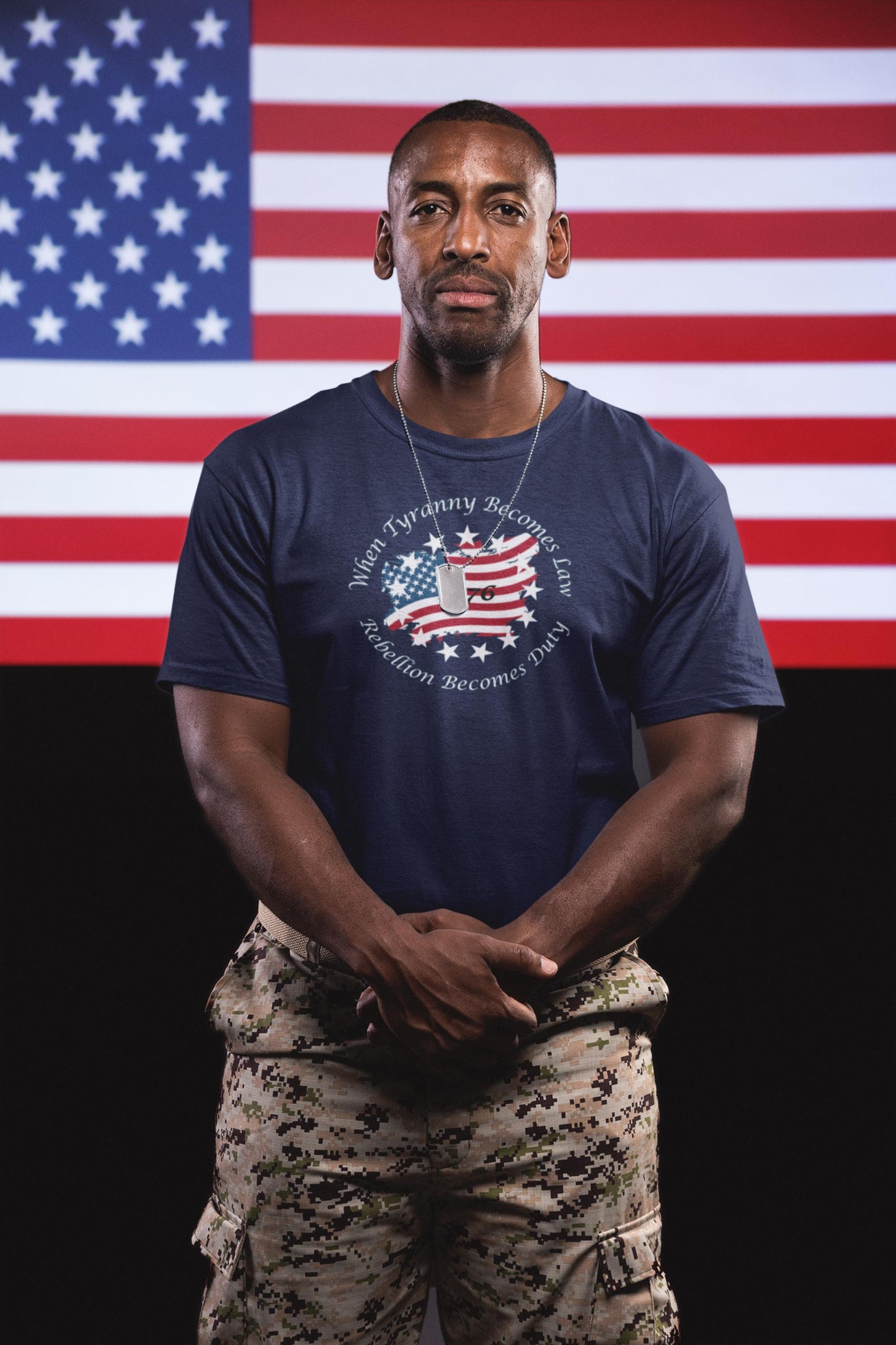 Veteran wearing a navy blue patriotic t-shirt with a distressed American flag design and the quote &quot;When Tyranny Becomes Law, Rebellion Becomes Duty,&quot; paired with digital camo pants—standing proudly in front of a large American flag backdrop.