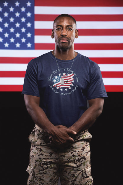 Veteran wearing a navy blue patriotic t-shirt with a distressed American flag design and the quote &quot;When Tyranny Becomes Law, Rebellion Becomes Duty,&quot; paired with digital camo pants—standing proudly in front of a large American flag backdrop.