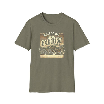 An olive green unisex t-shirt displayed flat on a white background, featuring a rustic graphic that reads &quot;Raised on Country Roads.&quot; The vintage-style design showcases a countryside scene with a classic pickup truck driving along a winding road past a barn, pine trees, and mountains, all under a rising sun. The artwork uses earthy tones and retro shading to evoke a nostalgic, rural Americana vibe.