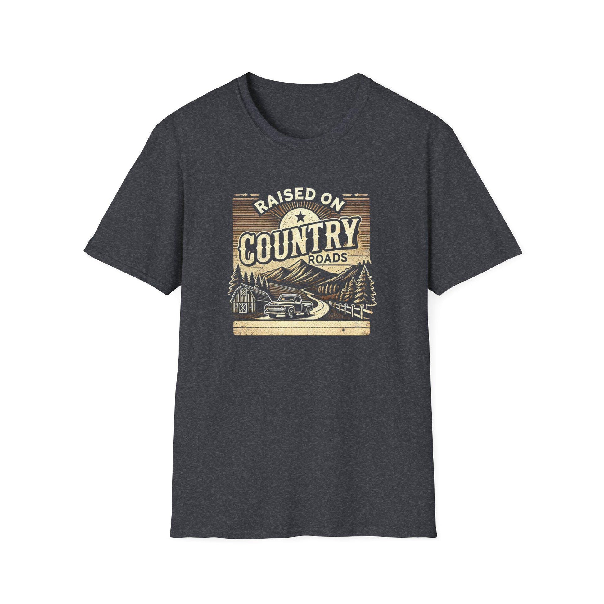 A dark heather gray unisex t-shirt is displayed flat on a white background, featuring a vintage-style graphic with the text &quot;Raised on Country Roads.&quot; The artwork shows a classic truck driving along a winding road through a rural landscape with pine trees, a barn, and distant mountains under a rising sun. The design uses muted tan, cream, and brown tones to create a nostalgic, rustic country feel.