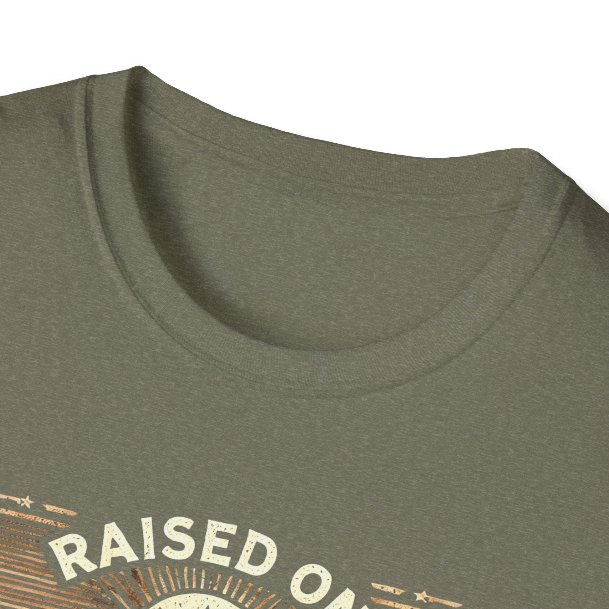 Close-up image of the neckline of an olive green t-shirt, showing a soft crew neck seam and part of a printed design that reads &quot;RAISED ON&quot; in off-white, vintage-style lettering. The fabric has a slightly heathered texture, and the partial graphic reveals sun rays and star accents in warm tones, suggesting a rustic or retro theme.