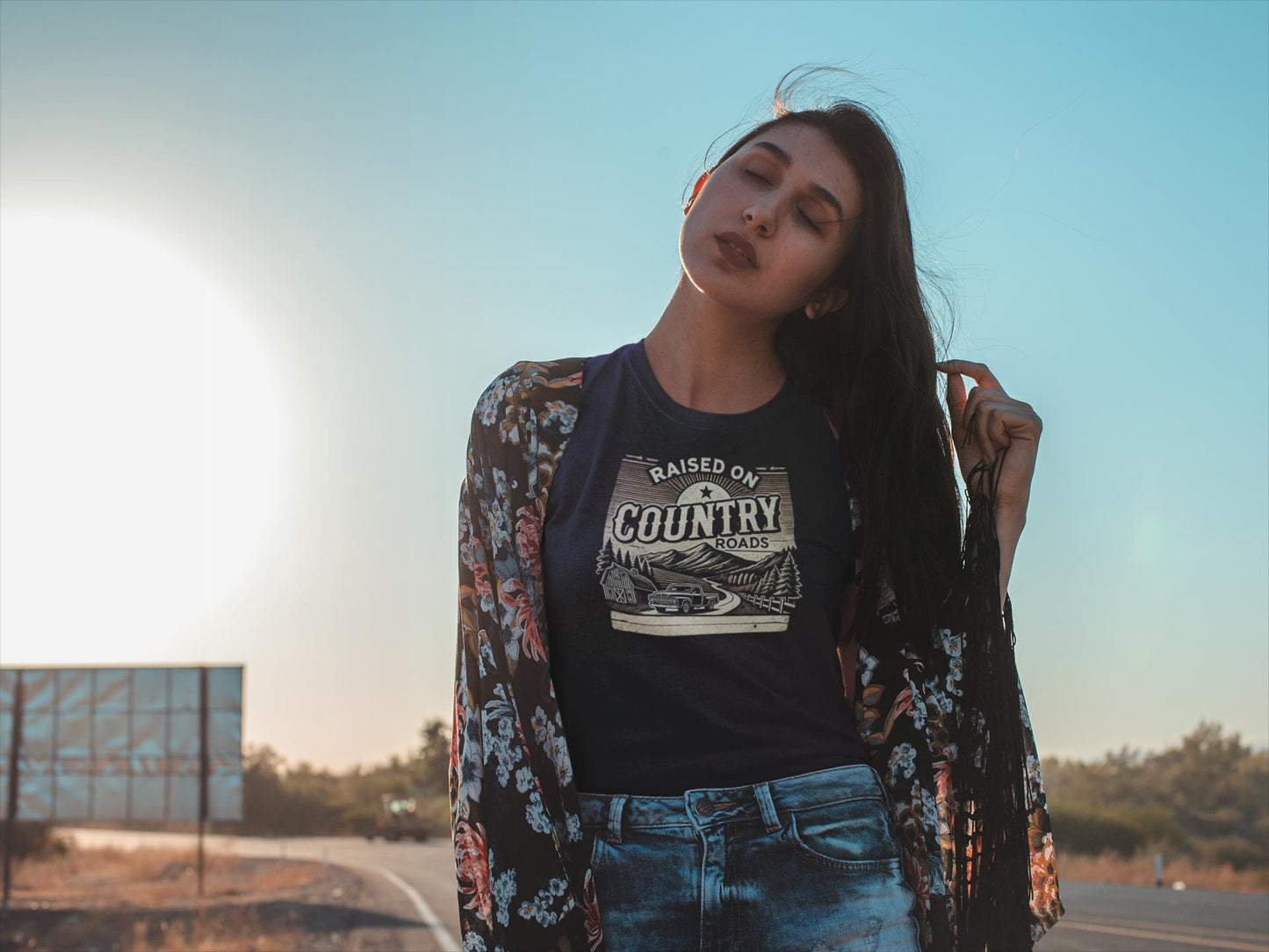 A woman stands by a rural roadside at sunset, eyes closed and head tilted slightly as she holds a strand of her long dark hair. She wears a navy blue t-shirt that says &quot;Raised on Country Roads&quot; with a retro-style graphic of a scenic drive past a barn and pine trees, along with high-waisted denim jeans and a floral kimono with fringe. The bright sun and clear blue sky create a warm, serene atmosphere in the background.