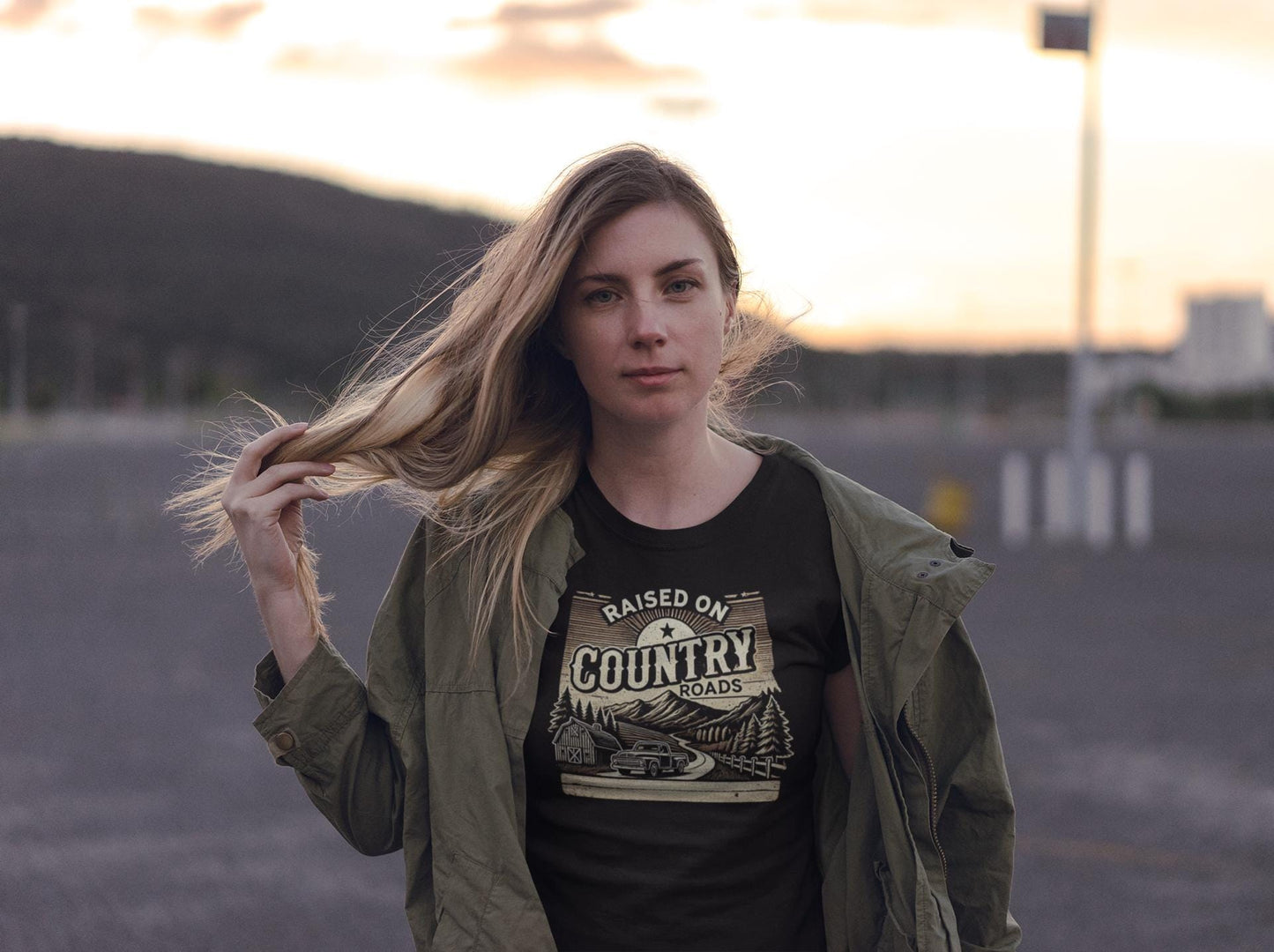 A woman with long blonde hair stands outdoors at sunset, gently holding her hair with one hand. She wears an olive green jacket over a black t-shirt that reads &quot;Raised on Country Roads&quot; with a vintage-style graphic of a car driving through a rural landscape with trees, a barn, and mountains. The background features a mountainous horizon and a mostly empty parking lot, evoking a peaceful, rustic atmosphere.