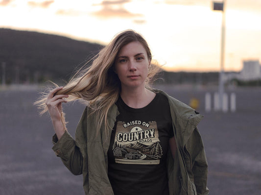 A woman with long blonde hair stands outdoors at sunset, gently holding her hair with one hand. She wears an olive green jacket over a black t-shirt that reads &quot;Raised on Country Roads&quot; with a vintage-style graphic of a car driving through a rural landscape with trees, a barn, and mountains. The background features a mountainous horizon and a mostly empty parking lot, evoking a peaceful, rustic atmosphere.