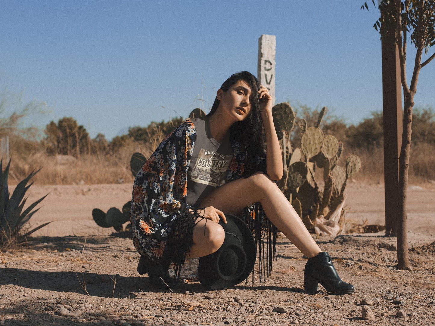 A woman poses in a desert setting, crouched on one knee beside a cactus and holding a black wide-brimmed hat. She wears a gray t-shirt with the text &quot;Raised on Country Roads&quot; and a vintage-style countryside graphic, paired with a floral kimono, black ankle boots, and long black fringe accents. The background features dry brush, agave plants, and a wooden post with a weathered sign, all under a clear blue sky. The overall vibe is rustic, boho, and Western-inspired.