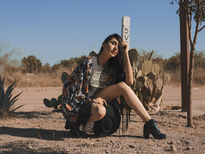 A woman poses in a desert setting, crouched on one knee beside a cactus and holding a black wide-brimmed hat. She wears a gray t-shirt with the text &quot;Raised on Country Roads&quot; and a vintage-style countryside graphic, paired with a floral kimono, black ankle boots, and long black fringe accents. The background features dry brush, agave plants, and a wooden post with a weathered sign, all under a clear blue sky. The overall vibe is rustic, boho, and Western-inspired.