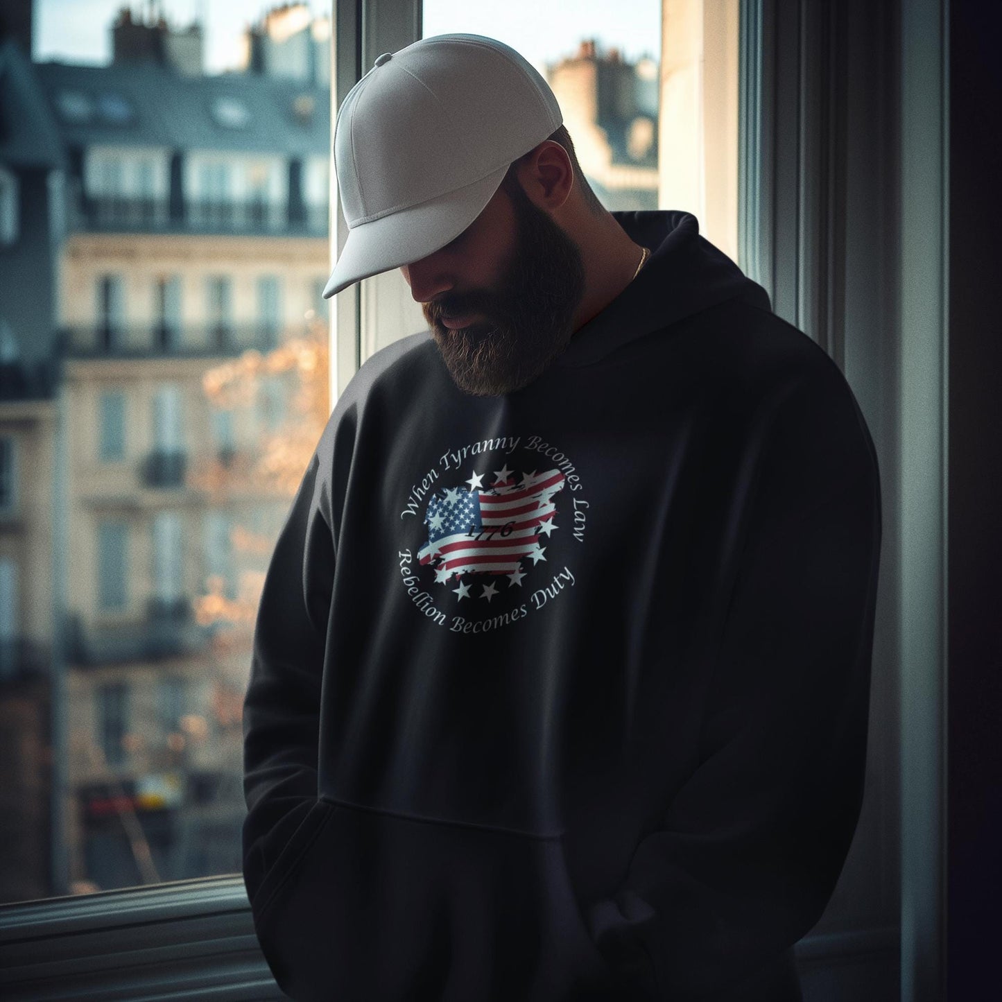 Bearded man wearing black patriotic hoodie with distressed American flag and “When Tyranny Becomes Law, Rebellion Becomes Duty” quote, standing by a window with urban buildings in the background. 1776 unisex pullover sweatshirt for men, bold conservative statement apparel, pro-America fashion, ideal for political expression or patriotic gift ideas.