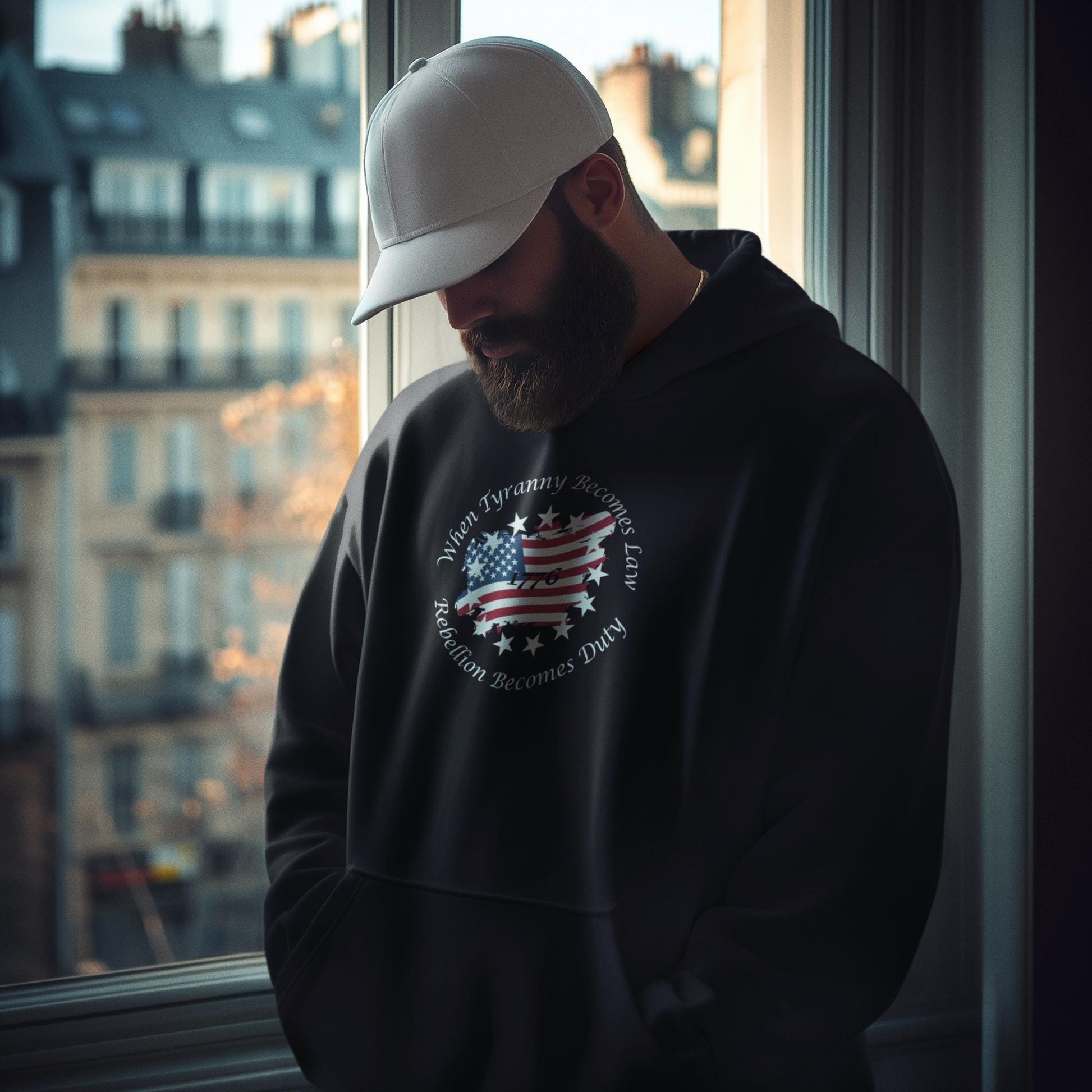 Bearded man wearing black patriotic hoodie with distressed American flag and “When Tyranny Becomes Law, Rebellion Becomes Duty” quote, standing by a window with urban buildings in the background. 1776 unisex pullover sweatshirt for men, bold conservative statement apparel, pro-America fashion, ideal for political expression or patriotic gift ideas.