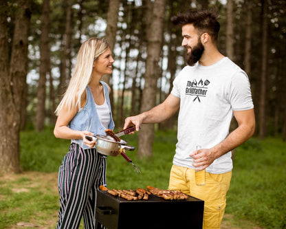 Man wearing a white T-shirt with the phrase “The Grillfather” and grilling icons, smiling while barbecuing outdoors with a woman holding a pot.