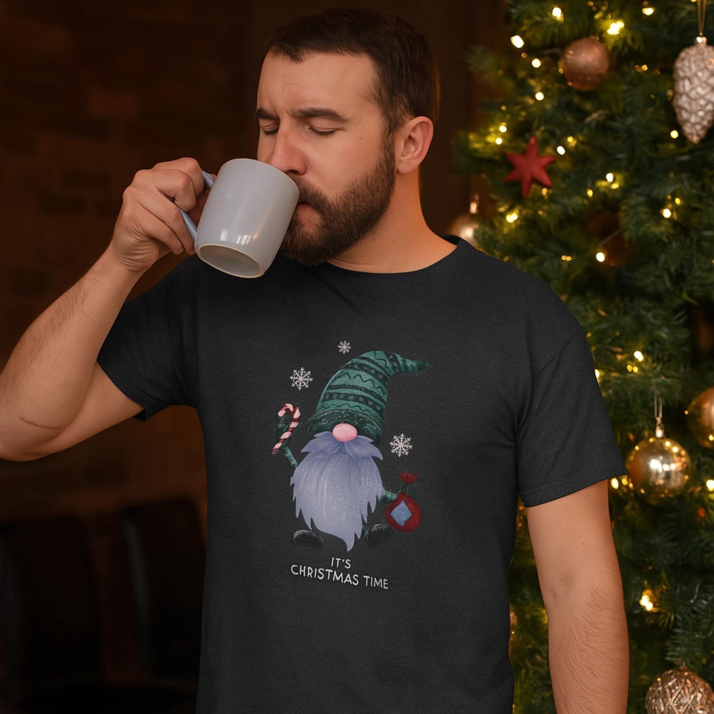 A man stands beside a decorated Christmas tree, sipping from a light gray mug with his eyes closed in a cozy, relaxed moment. He is wearing a black T-shirt featuring a festive gnome holding a candy cane and ornament, surrounded by snowflakes, with the text &quot;It&#39;s Christmas Time&quot; printed below the graphic. The warm lighting and holiday décor create a peaceful, festive ambiance.