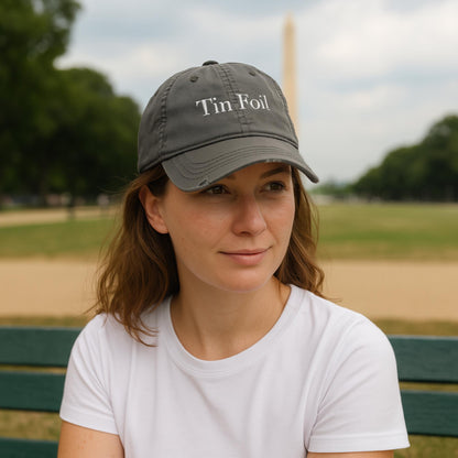 A woman wearing a gray baseball cap with the words &quot;Tin Foil&quot; embroidered in white sits on a green bench in a park. She has a calm expression and is dressed in a plain white t-shirt. In the blurred background, the Washington Monument is visible, suggesting a location in Washington, D.C., with green trees and an overcast sky.