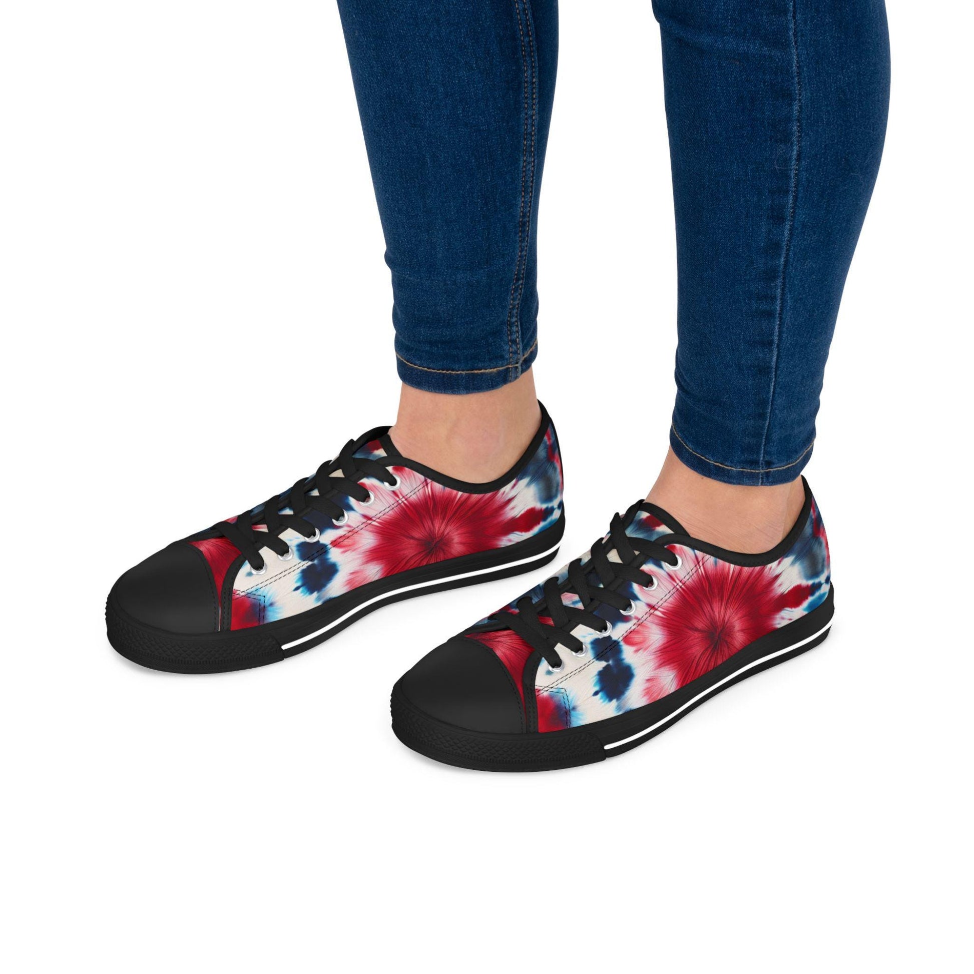 Person wearing a pair of low-top canvas sneakers with a bold red, white, and blue tie-dye design. The shoes feature black laces, black rubber soles, and black toe caps, creating a striking contrast. The person is dressed in dark blue skinny jeans, and the image showcases the vibrant patriotic shoes from a side angle, perfect for casual, festive occasions.