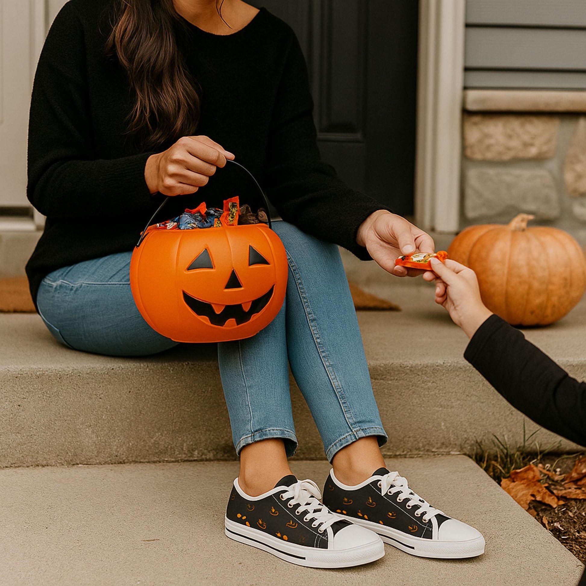 Women&#39;s Halloween low-top canvas sneakers with orange jack-o&#39;-lantern face design, black and white lace-up shoes perfect for spooky season outfits and trick-or-treat night – shown on a woman handing out candy with a pumpkin candy bucket on a front porch decorated for fall.