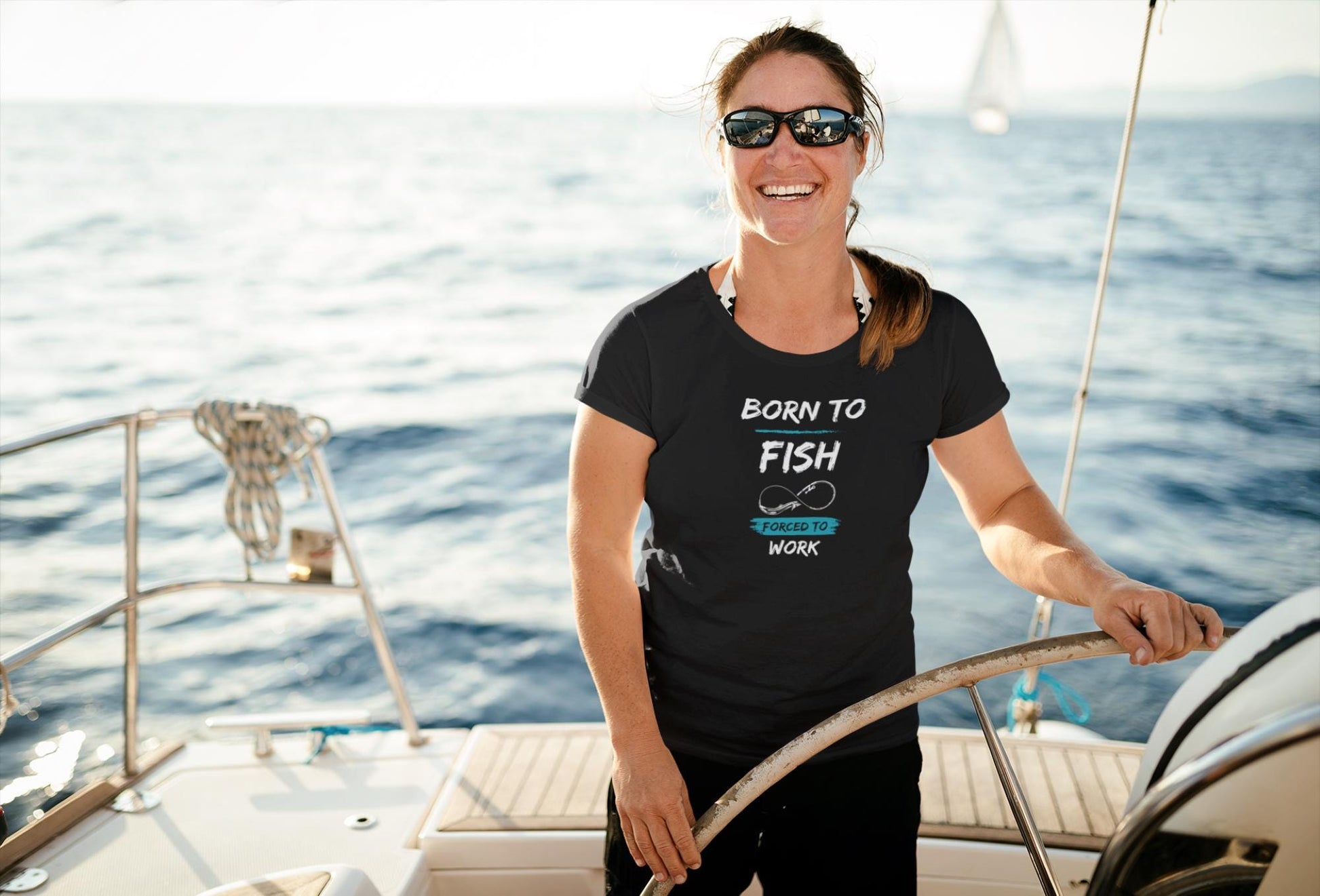 Happy woman steering a sailboat on the open water, wearing sunglasses and a black t-shirt that reads &#39;Born to Fish, Forced to Work&#39; in white and teal text with a fishing hook graphic. The sun is shining brightly over the calm sea, creating a relaxed and adventurous boating scene.