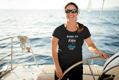 Happy woman steering a sailboat on the open water, wearing sunglasses and a black t-shirt that reads &#39;Born to Fish, Forced to Work&#39; in white and teal text with a fishing hook graphic. The sun is shining brightly over the calm sea, creating a relaxed and adventurous boating scene.