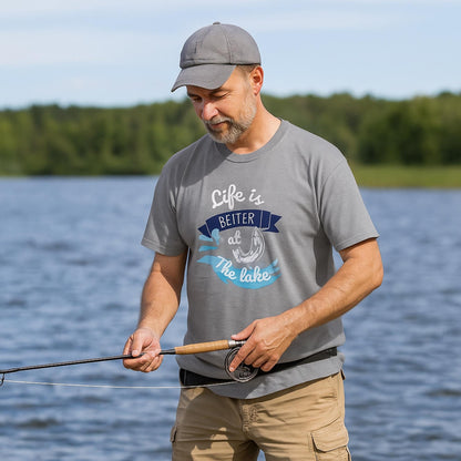 Man fishing by a lake while wearing a gray t-shirt with a colorful graphic that reads &#39;Life is Better at the Lake,&#39; featuring a fish hook, water splash, and banner elements in blue and white. He is holding a fishing rod and wearing a gray cap and beige cargo shorts, with trees and water in the background.