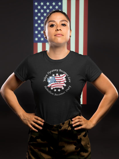 Confident woman standing with hands on hips, wearing a black fitted t-shirt featuring a distressed American flag in the shape of the U.S., surrounded by stars and the quote &#39;When Tyranny Becomes Law, Rebellion Becomes Duty - 1776.&#39; She poses in front of a large vertical American flag backdrop, wearing camouflage pants.