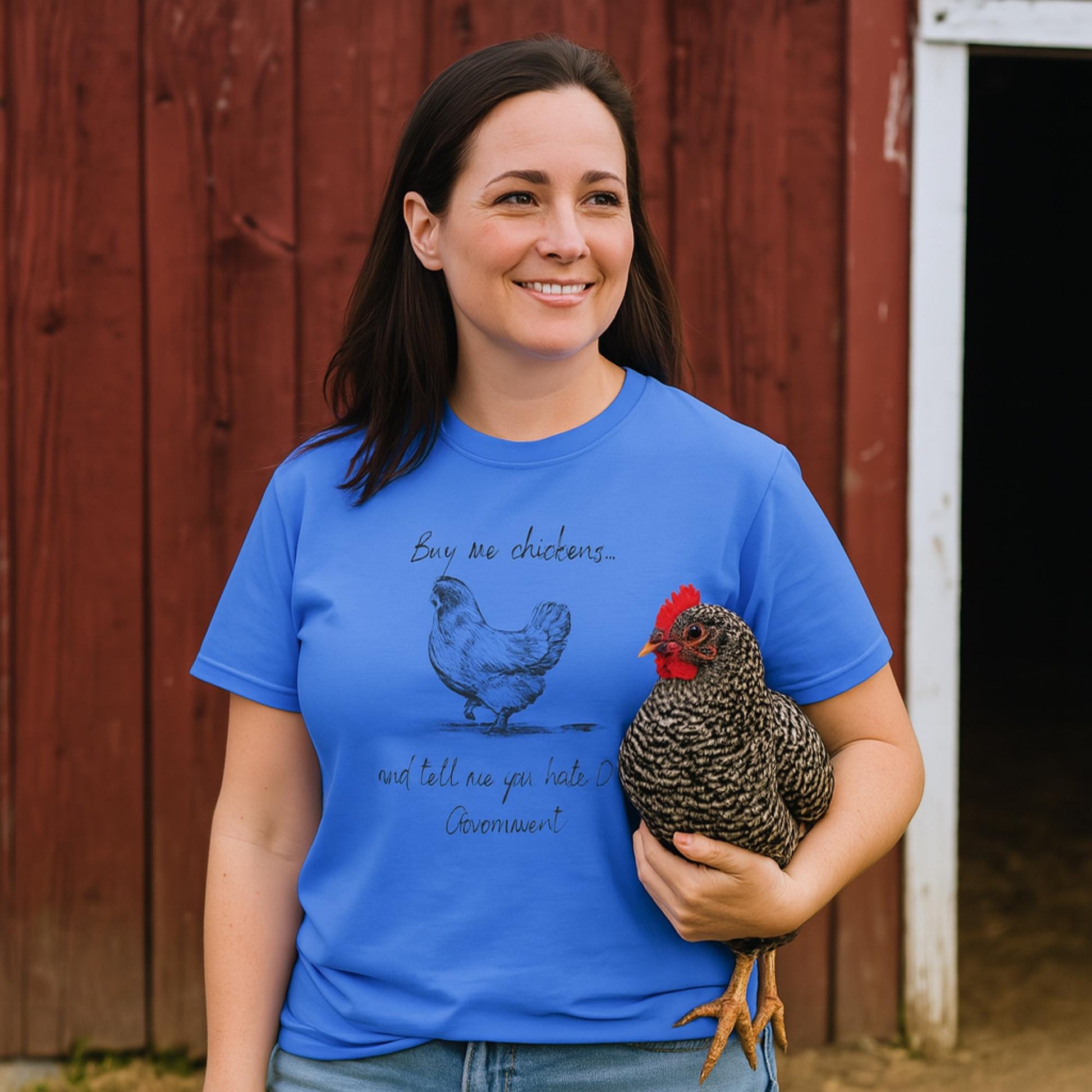 A woman stands in front of a red barn, smiling and holding a black-and-white speckled chicken. She is wearing a bright blue T-shirt with a hand-drawn illustration of a chicken and the humorous text: &quot;Buy me chickens... and tell me you hate the government.