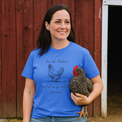 A woman stands in front of a red barn, smiling and holding a black-and-white speckled chicken. She is wearing a bright blue T-shirt with a hand-drawn illustration of a chicken and the humorous text: &quot;Buy me chickens... and tell me you hate the government.