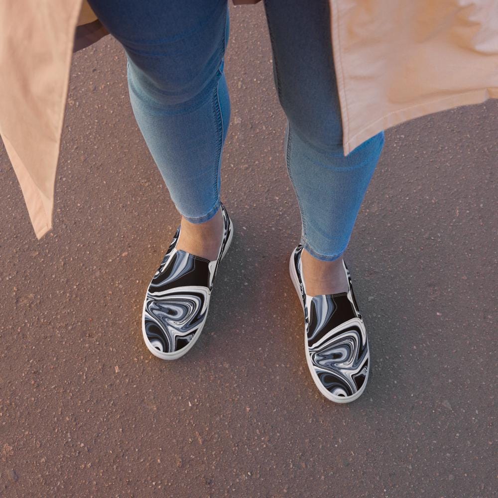 Close-up view of a woman wearing black and white marbled abstract slip-on canvas shoes, styled with light blue skinny jeans and a beige trench coat, standing on an asphalt street. Modern casual footwear for women, perfect for urban walking style.