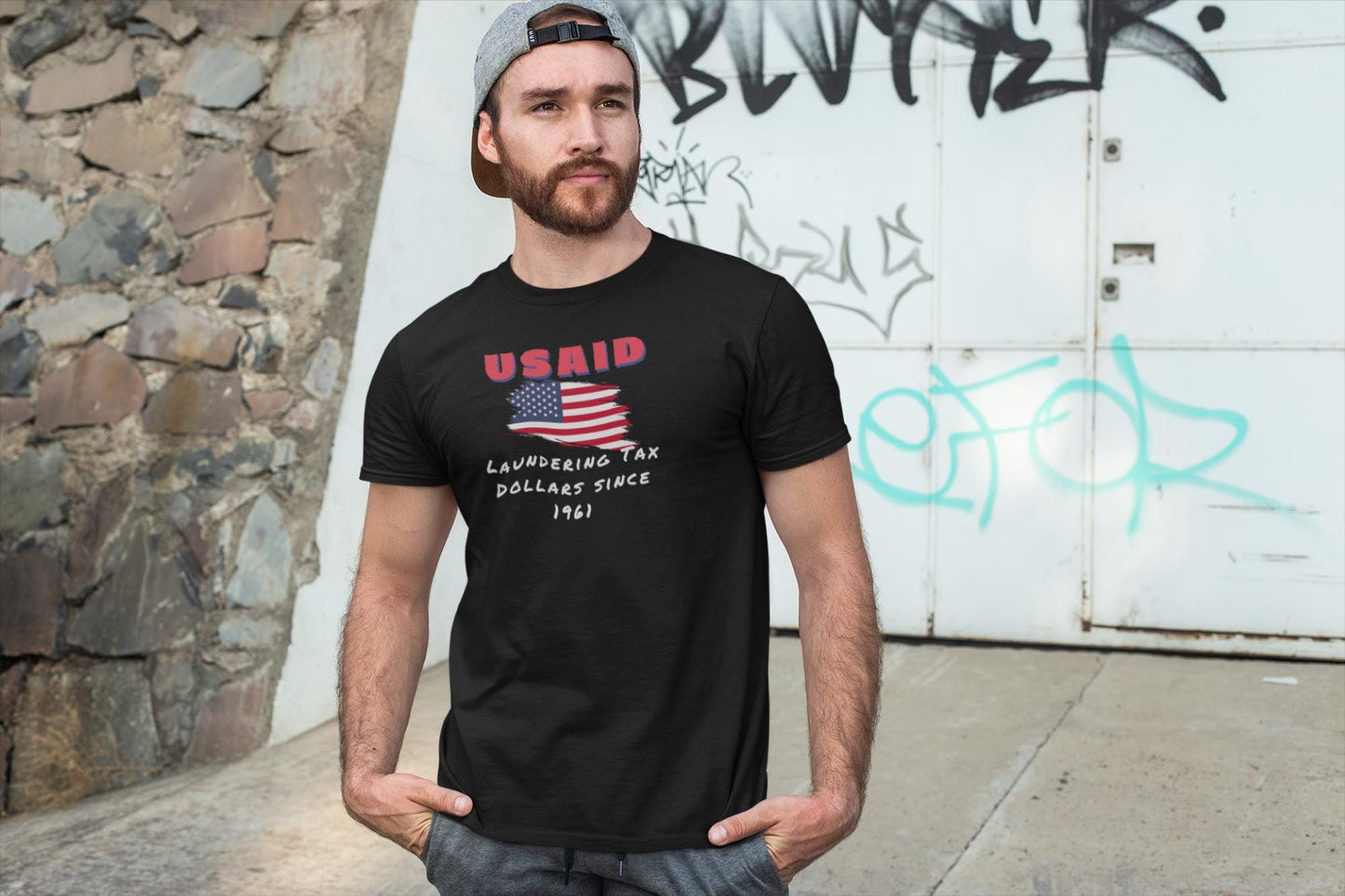 A man stands casually outdoors in front of a graffiti-covered wall and stone structure, with his hands in his pockets. He is wearing a backward gray cap, athletic shorts, and a black T-shirt featuring a distressed American flag graphic. Above the flag, red text reads &quot;USAID,&quot; and below it, white handwritten-style text says: &quot;Laundering Tax Dollars Since 1961.&quot; The shirt has a satirical political tone, mimicking a vintage protest design.