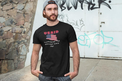 A man stands casually outdoors in front of a graffiti-covered wall and stone structure, with his hands in his pockets. He is wearing a backward gray cap, athletic shorts, and a black T-shirt featuring a distressed American flag graphic. Above the flag, red text reads &quot;USAID,&quot; and below it, white handwritten-style text says: &quot;Laundering Tax Dollars Since 1961.&quot; The shirt has a satirical political tone, mimicking a vintage protest design.