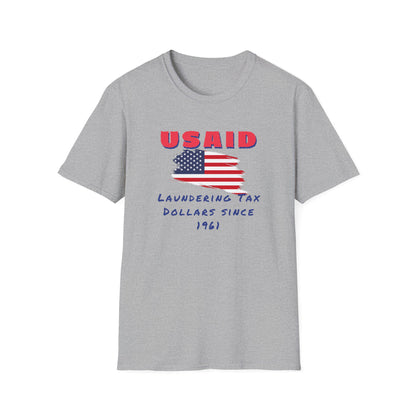 A heather gray T-shirt is displayed flat against a white background. The front of the shirt features a distressed American flag graphic in the center. Above the flag, bold red letters spell out &quot;USAID.&quot; Below the flag, blue handwritten-style text reads: &quot;Laundering Tax Dollars Since 1961.&quot; The design conveys a satirical political message with a protest-style aesthetic.