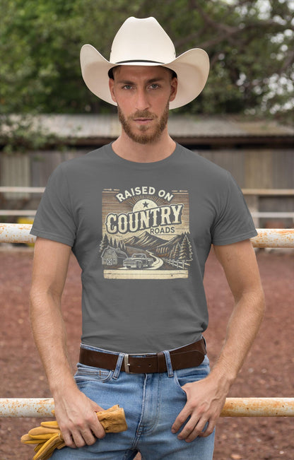 A man stands confidently in front of a rustic barnyard fence, wearing a white cowboy hat and a gray t-shirt that reads &quot;Raised on Country Roads&quot; with a vintage-style design of a rural landscape, including a barn, trees, mountains, and a classic truck. He holds a pair of work gloves in one hand and rests the other on his hip. His outfit includes light blue jeans and a brown leather belt, embodying a classic Western, country lifestyle look.