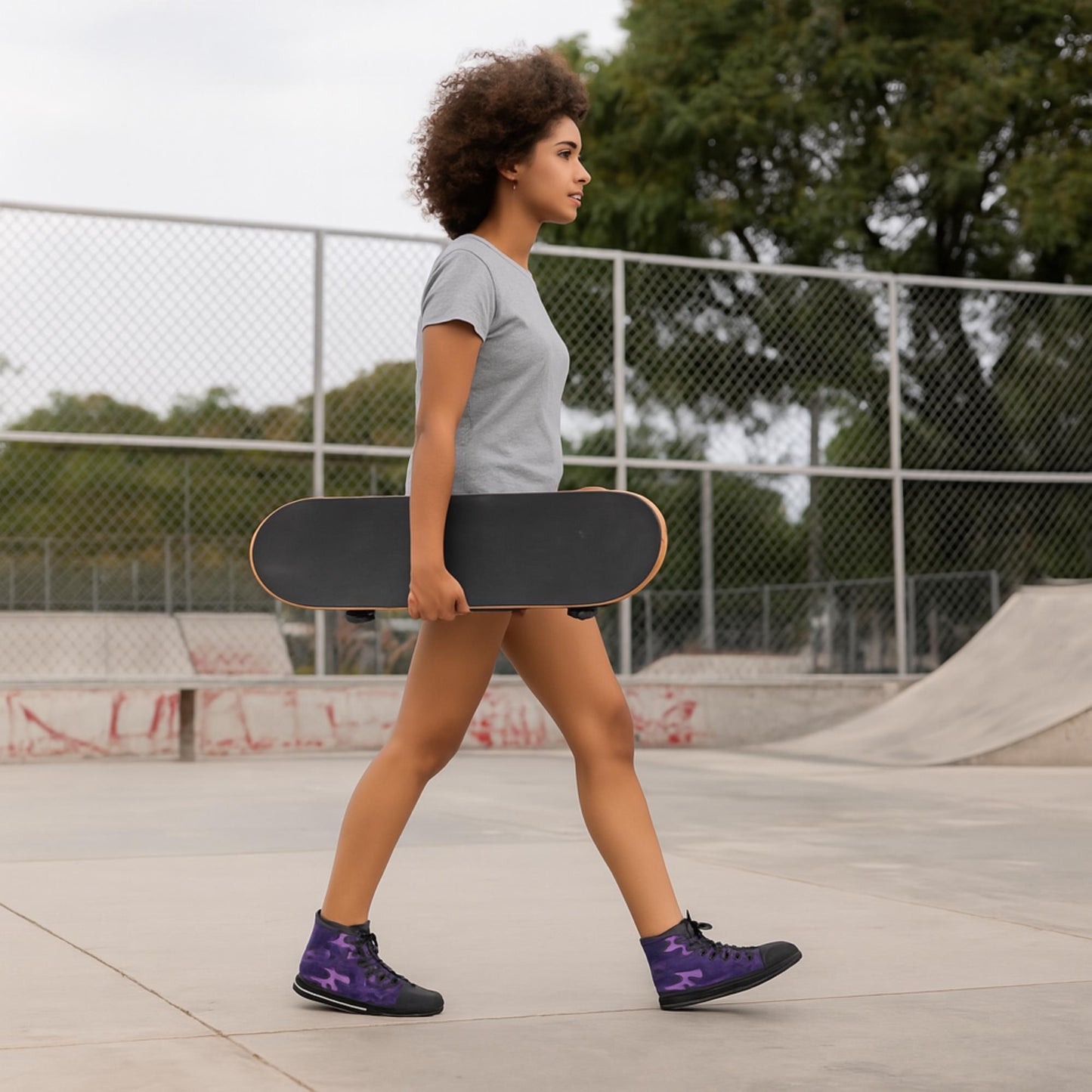 A young woman with curly hair is walking through a skatepark while holding a black skateboard by her side. She is wearing a light gray t-shirt, shorts, and purple camouflage high-top sneakers with black laces and soles. A chain-link fence and skate ramps are visible in the background, along with trees.
