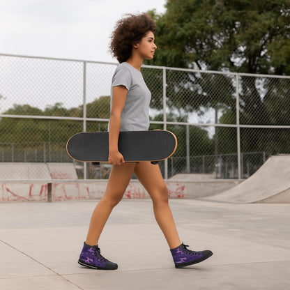 A young woman with curly hair is walking through a skatepark while holding a black skateboard by her side. She is wearing a light gray t-shirt, shorts, and purple camouflage high-top sneakers with black laces and soles. A chain-link fence and skate ramps are visible in the background, along with trees.