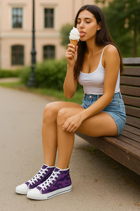 A young woman with long dark hair is sitting on a wooden bench in a park, wearing a white tank top, high-waisted denim shorts, and purple camouflage high-top sneakers with white laces. She is holding a vanilla ice cream cone and playfully sticking out her tongue to taste it, with her eyes closed. Trees and a light-colored building are visible in the background.