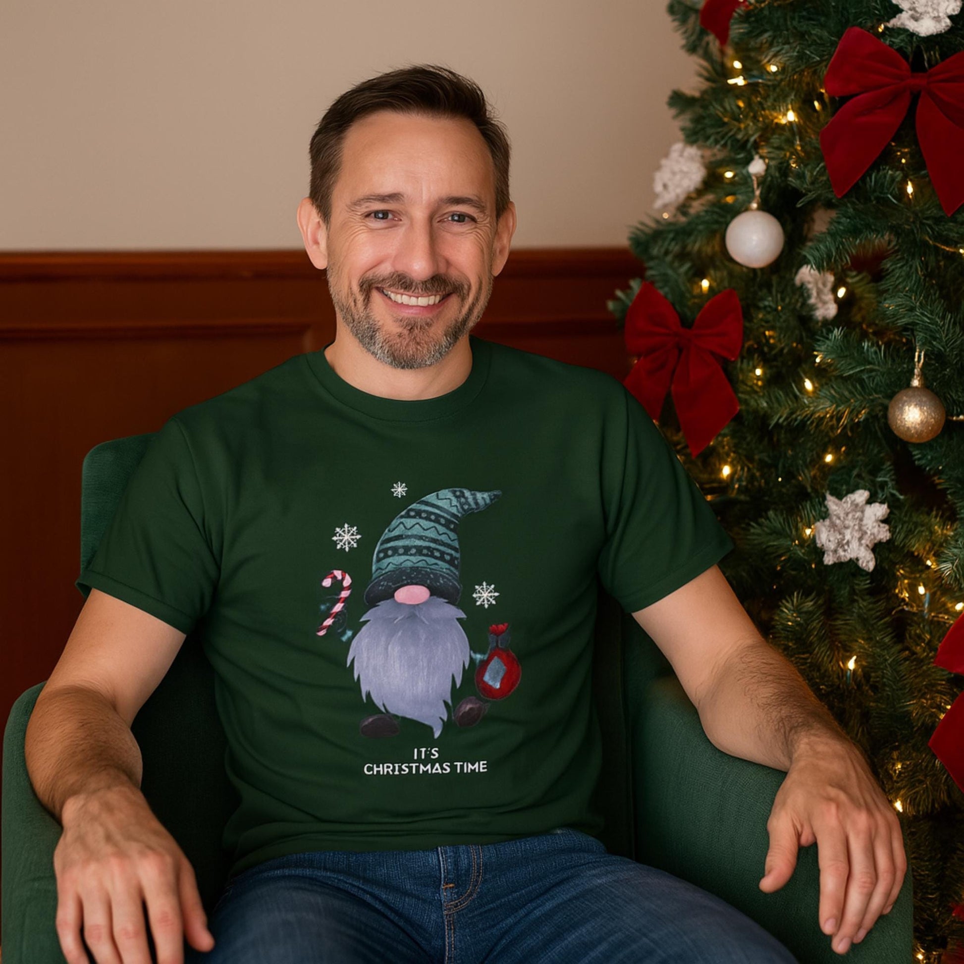 A man sits smiling in a green armchair next to a decorated Christmas tree adorned with red bows and ornaments. He is wearing a dark green T-shirt featuring a festive gnome graphic holding a candy cane and an ornament, with snowflakes around and the text &quot;It&#39;s Christmas Time&quot; printed below. The setting is warm and festive, evoking a cheerful holiday atmosphere.
