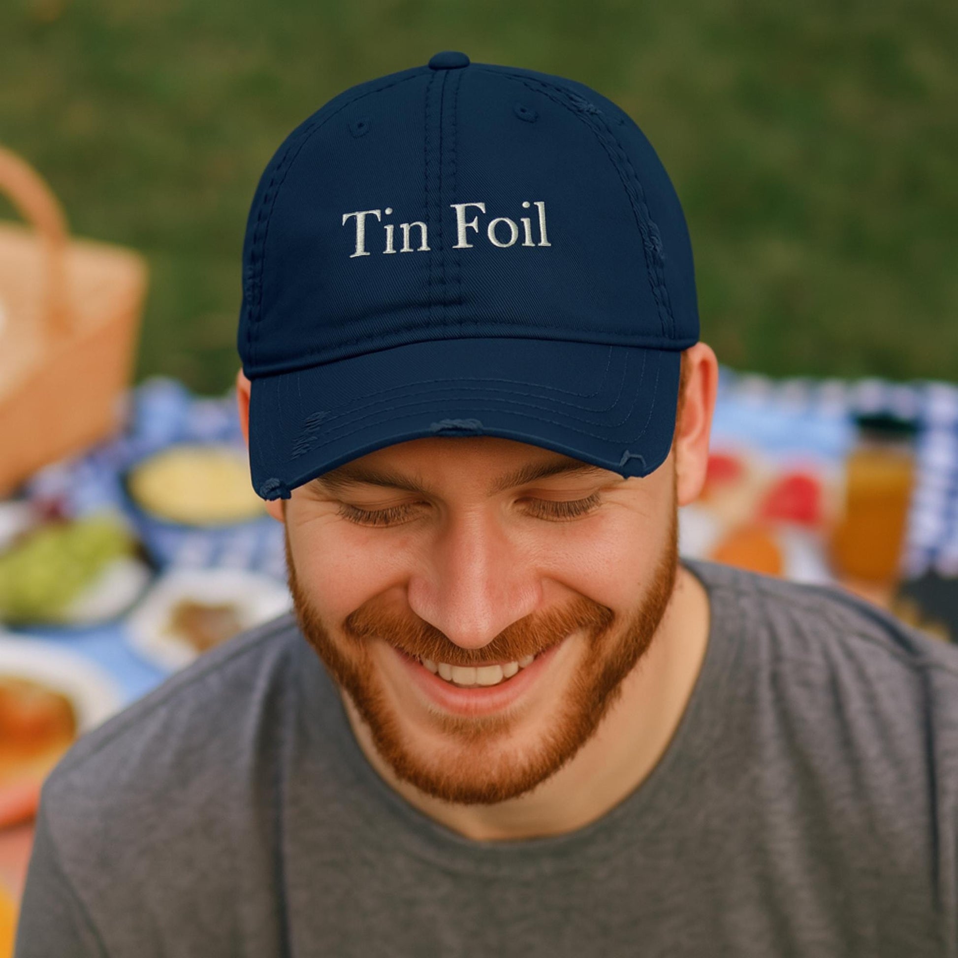 A smiling man with a red beard wears a navy blue baseball cap embroidered with the words &quot;Tin Foil&quot; in white. He is seated outdoors at a picnic, with a blurred background showing a checkered blanket, food, and a wicker picnic basket on the grass. He is dressed casually in a gray t-shirt and appears relaxed and happy.