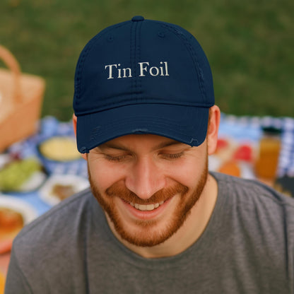 A smiling man with a red beard wears a navy blue baseball cap embroidered with the words &quot;Tin Foil&quot; in white. He is seated outdoors at a picnic, with a blurred background showing a checkered blanket, food, and a wicker picnic basket on the grass. He is dressed casually in a gray t-shirt and appears relaxed and happy.