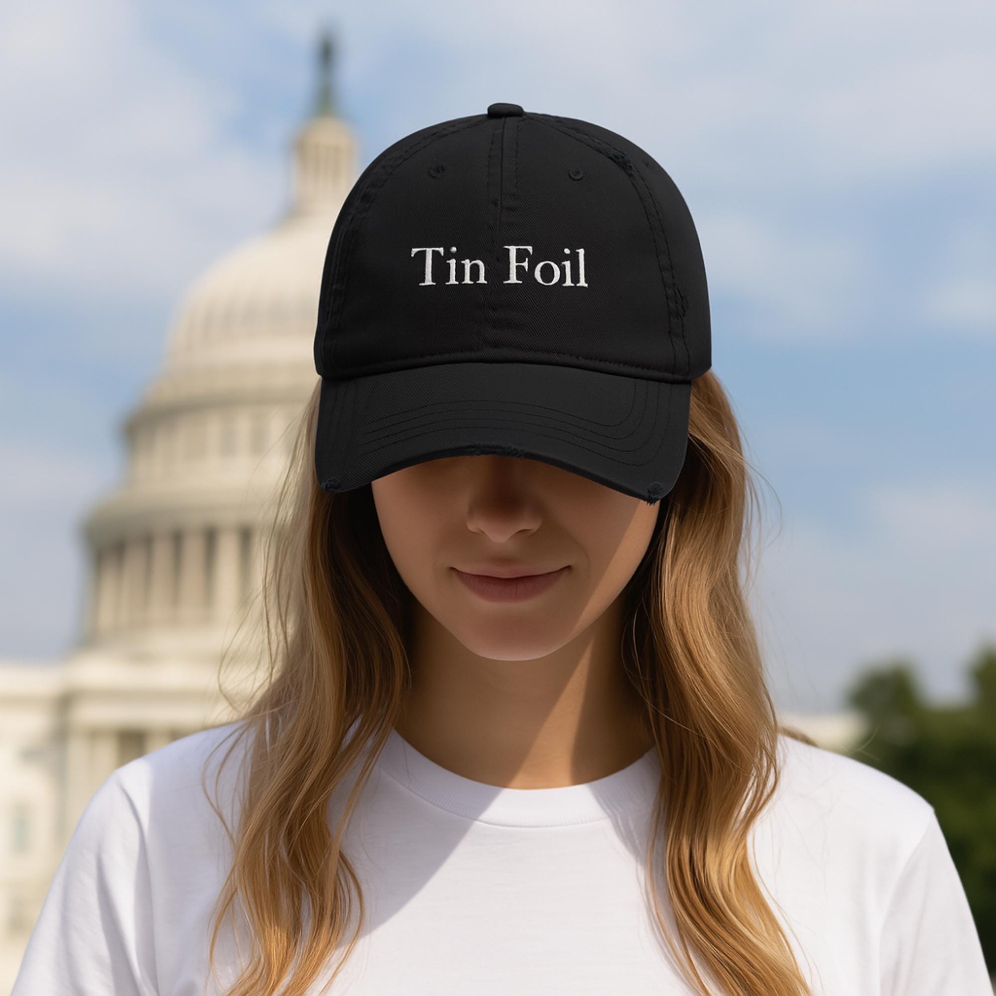 A young woman wearing a black baseball cap with the words &quot;Tin Foil&quot; embroidered in white on the front. She stands in front of the U.S. Capitol building, slightly smiling with her head tilted down, and wears a plain white t-shirt. The background is softly blurred, emphasizing the cap and her face.