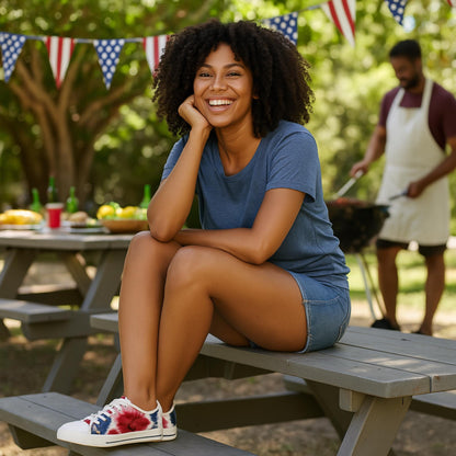 A young woman with curly hair sits on a picnic table bench outdoors, smiling brightly at the camera. She is wearing a casual blue t-shirt, denim shorts, and patriotic tie-dye sneakers. Behind her, a man in an apron grills food while red, white, and blue American flag bunting decorates the background, suggesting a festive summer barbecue or Fourth of July celebration.