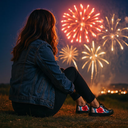 A woman sits on the grass at night, watching a vibrant fireworks display light up the sky. She is seen from behind, wearing a denim jacket, black leggings, and patriotic tie-dye sneakers. The scene captures a festive atmosphere, likely during a Fourth of July celebration, with glowing bursts of red and gold fireworks illuminating the dark evening.