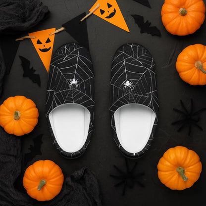 A pair of black slippers with white spiderweb and spider designs are displayed on a dark surface surrounded by mini orange pumpkins, black faux spiders, bat cutouts, black netting, and an orange and black jack-o&#39;-lantern banner. The Halloween-themed setup creates a festive and spooky vibe.