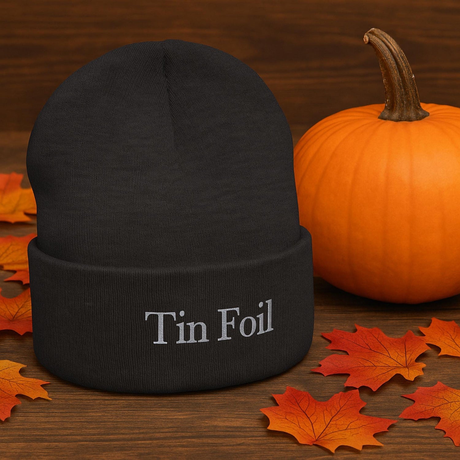 A black knit beanie with the words &quot;Tin Foil&quot; embroidered in silver thread is displayed on a wooden surface. Surrounding the beanie are scattered autumn leaves and a bright orange pumpkin, creating a cozy fall or Halloween-themed setting.