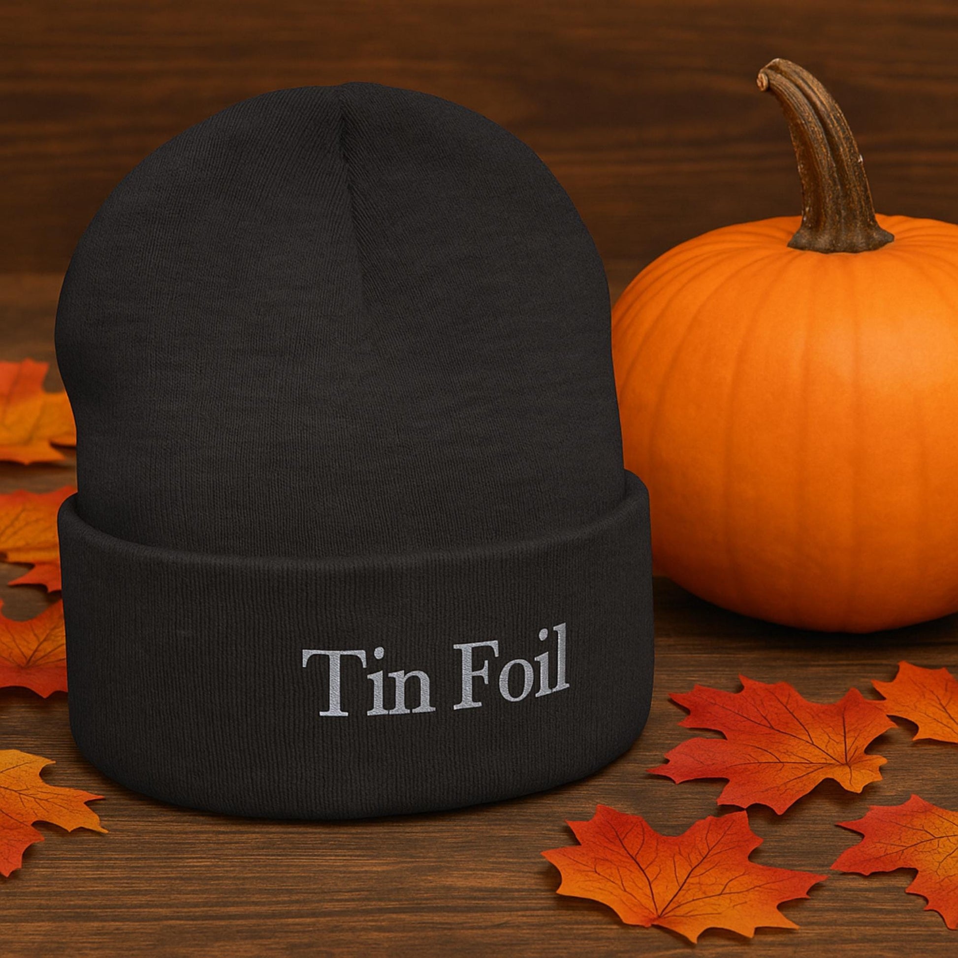A black knit beanie with the words &quot;Tin Foil&quot; embroidered in silver thread is displayed on a wooden surface. Surrounding the beanie are scattered autumn leaves and a bright orange pumpkin, creating a cozy fall or Halloween-themed setting.