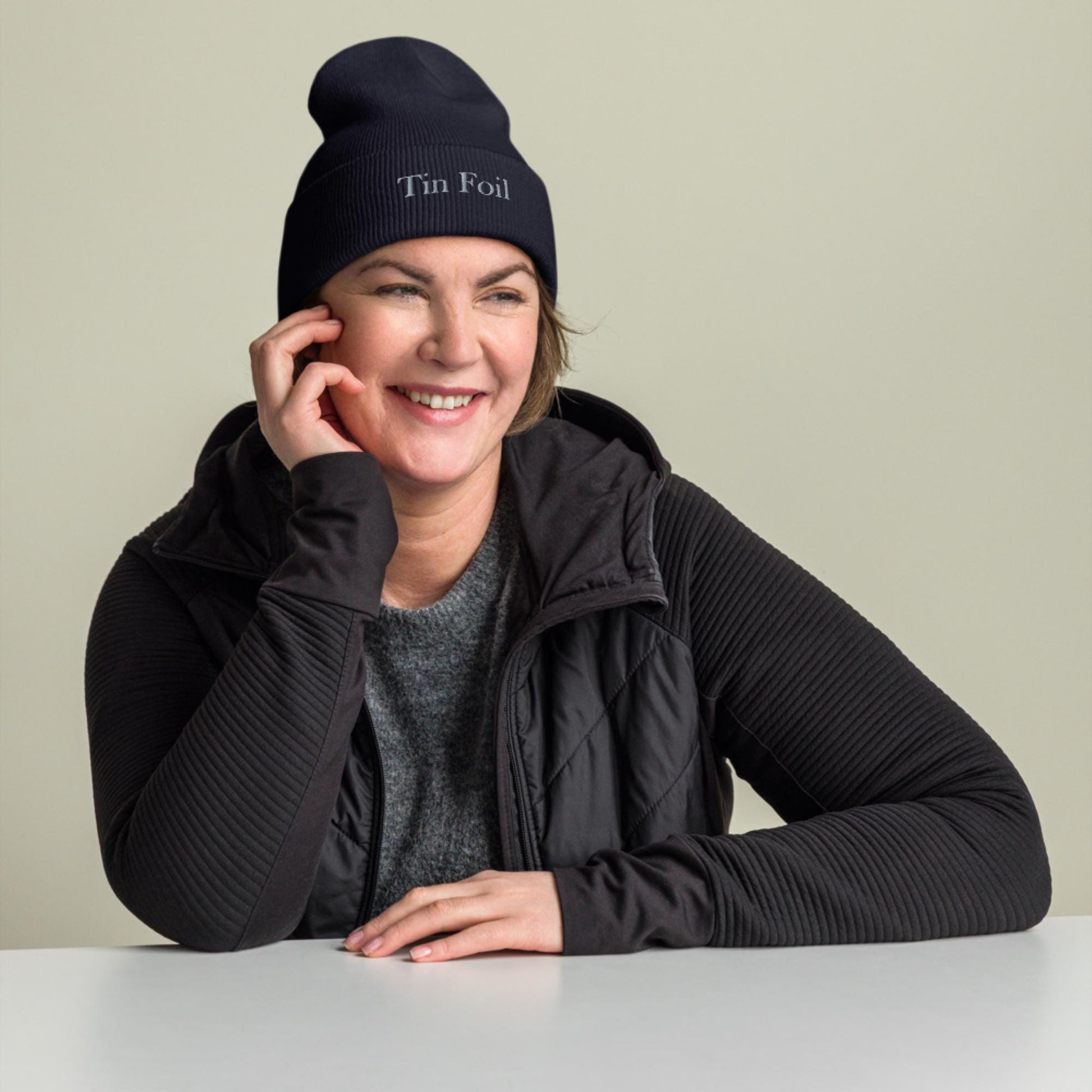 &quot;Smiling woman wearing a black beanie with the words &#39;Tin Foil&#39; embroidered on the front. She is dressed in a dark zip-up jacket over a gray top, resting her elbow on a white table and lightly touching her face, with a neutral beige background.&quot;