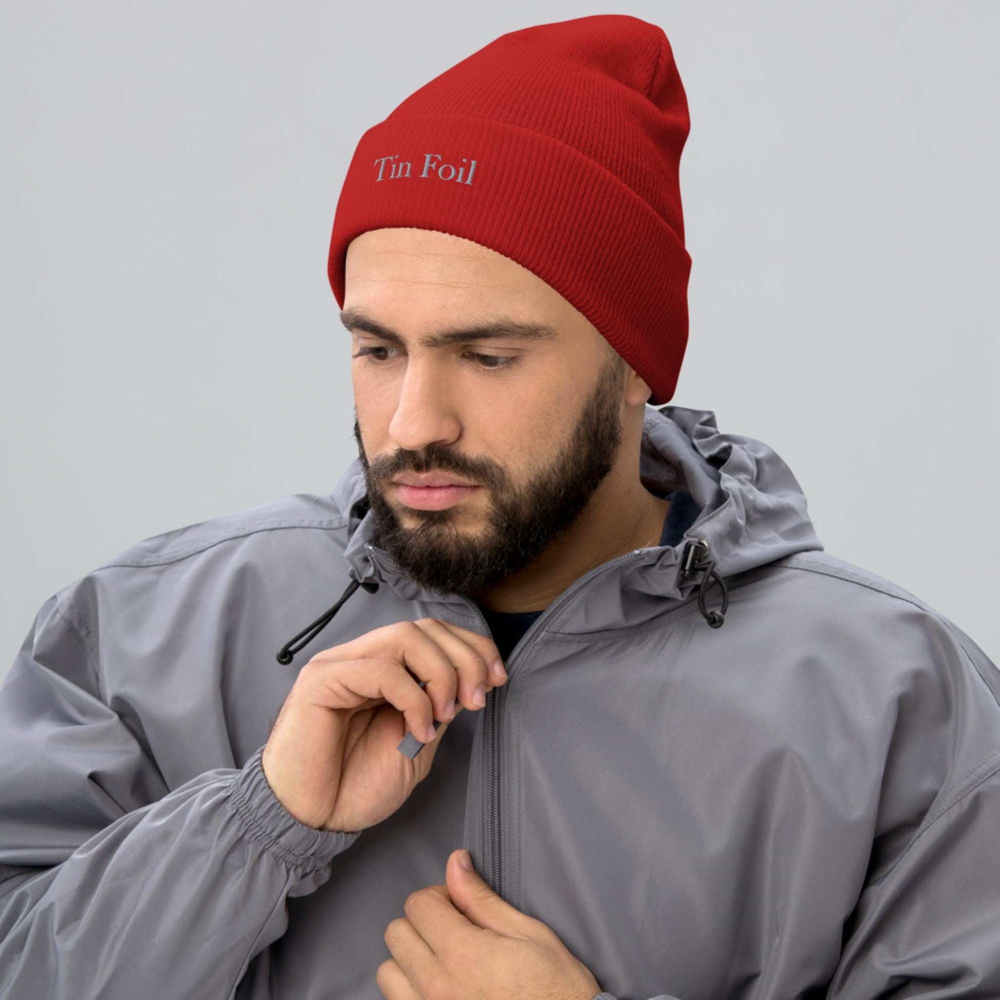 &quot;Man wearing a red beanie with the words &#39;Tin Foil&#39; embroidered on the front. He has a full beard and is dressed in a light gray hooded windbreaker, looking down while adjusting the zipper, against a plain gray background.&quot;