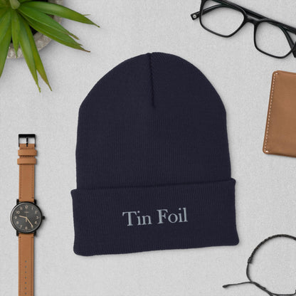 &quot;Flat lay of a navy blue beanie with the words &#39;Tin Foil&#39; embroidered on the front. The beanie is surrounded by accessories including a brown leather watch, black eyeglasses, a brown wallet, a black bracelet, and a small potted succulent, all arranged on a light gray surface.&quot;
