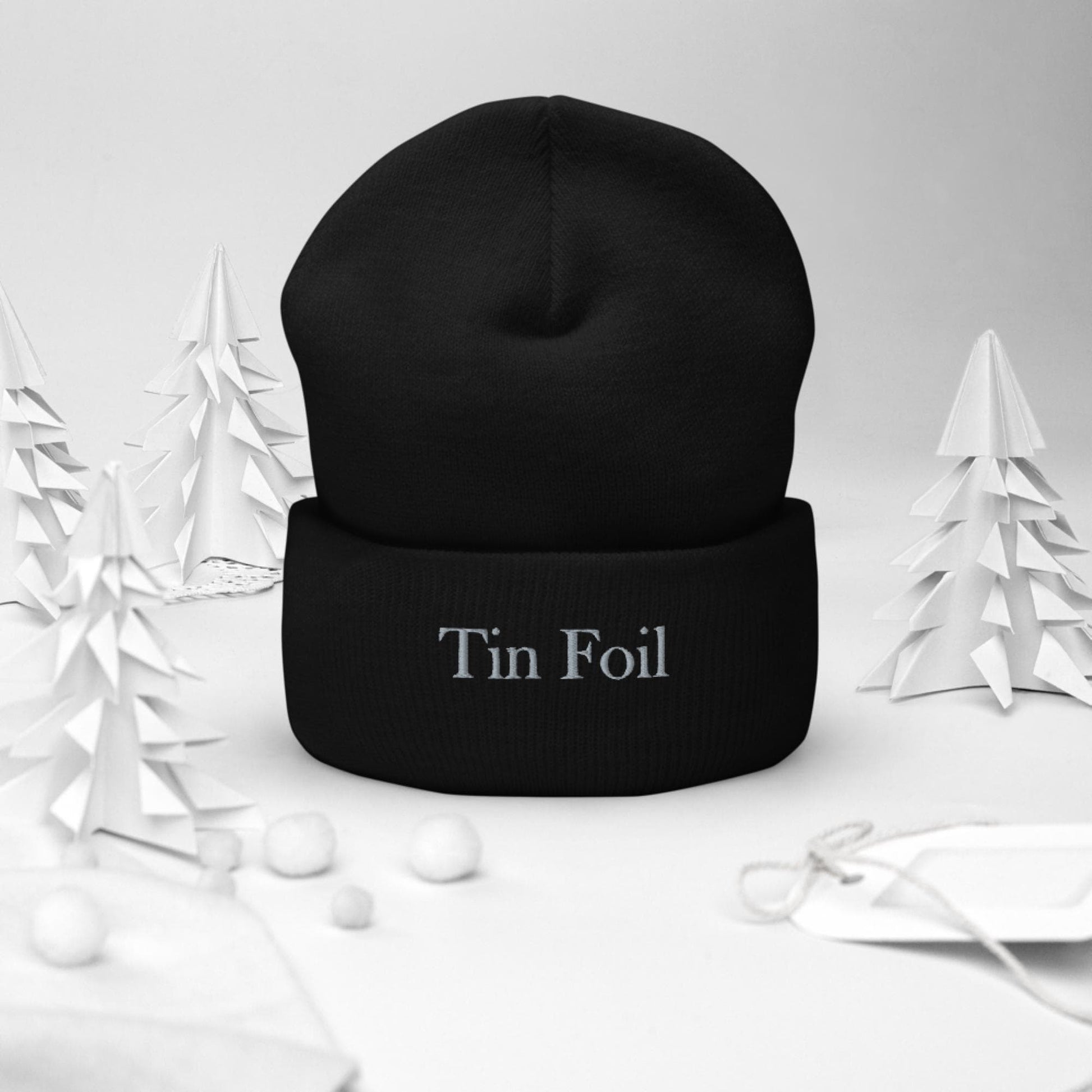 &quot;Black beanie with the words &#39;Tin Foil&#39; embroidered on the front, displayed upright in a winter-themed setting. Surrounding the beanie are white paper-cut trees, small white pom-pom snowballs, and a white gift tag with string, all set against a soft white background.&quot;