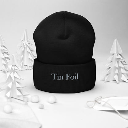 &quot;Black beanie with the words &#39;Tin Foil&#39; embroidered on the front, displayed upright in a winter-themed setting. Surrounding the beanie are white paper-cut trees, small white pom-pom snowballs, and a white gift tag with string, all set against a soft white background.&quot;