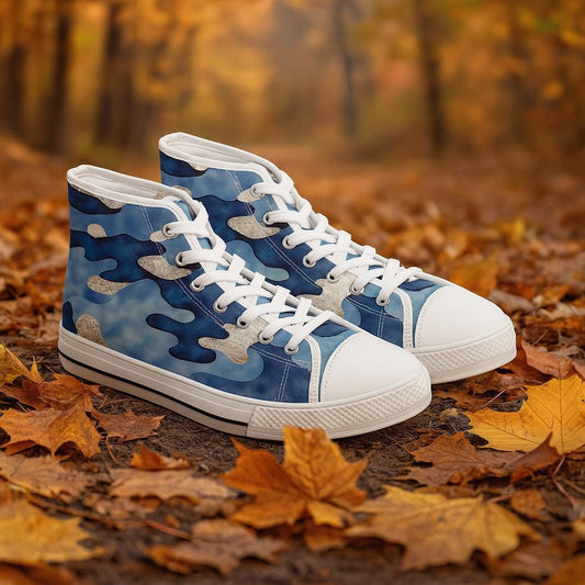Pair of high-top sneakers with a blue and gray camouflage pattern, featuring white laces and rubber soles. The shoes are placed on a forest floor covered with fallen autumn leaves, with a blurred background of trees in warm fall colors.
