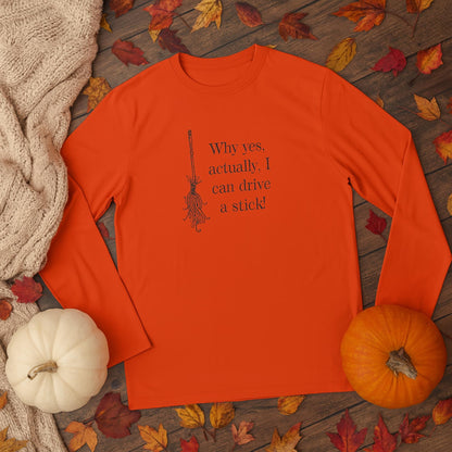 Orange long-sleeve shirt laid flat on a wooden surface surrounded by autumn leaves, a cozy beige knit sweater, a white pumpkin, and an orange pumpkin. The shirt features a black illustration of a broomstick and the humorous text: &#39;Why yes, actually, I can drive a stick!