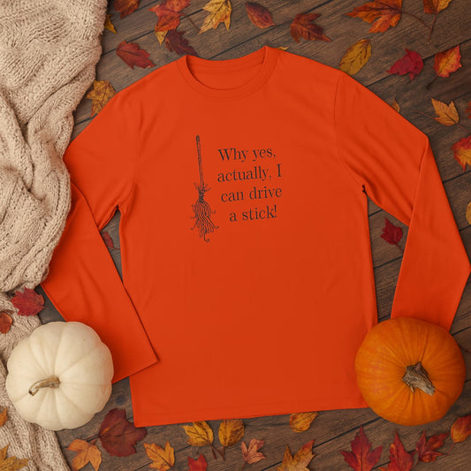 Orange long-sleeve shirt laid flat on a wooden surface surrounded by autumn leaves, a cozy beige knit sweater, a white pumpkin, and an orange pumpkin. The shirt features a black illustration of a broomstick and the humorous text: &#39;Why yes, actually, I can drive a stick!