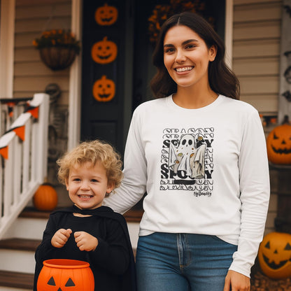Smiling woman wearing a white long-sleeve shirt with a Halloween ghost graphic and the word &#39;SEASON&#39; repeated in the background. She stands next to a young child in a black cape costume holding an orange pumpkin-shaped candy bucket. They are on a porch decorated with jack-o&#39;-lanterns and Halloween décor.