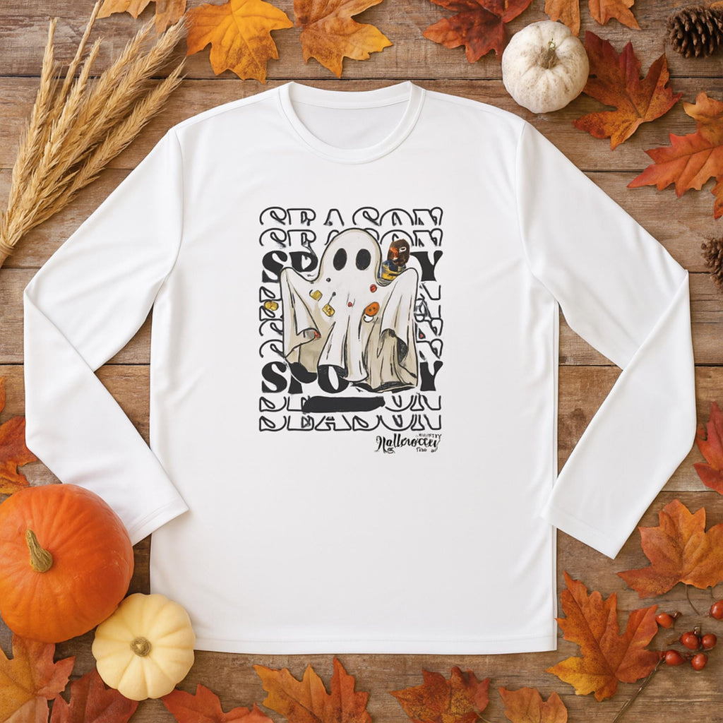 White long-sleeve shirt laid flat on a wooden surface surrounded by autumn leaves, pumpkins, pinecones, and dried wheat stalks. The shirt features a graphic of a ghost costume with candy and Halloween icons, set against a repeating words &#39;SEASON&#39; and &#39;SPOOKY&#39; text background. At the bottom, it says &#39;Halloween&#39; in stylized lettering.